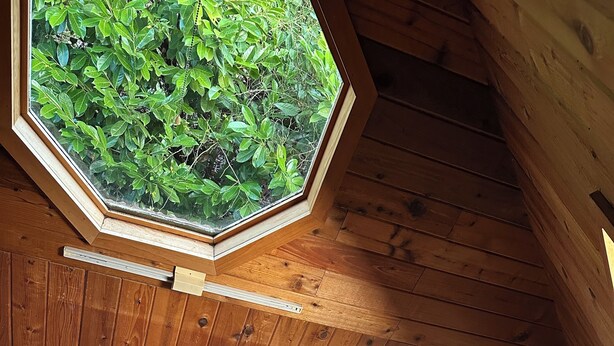 A cozy wooden cabin interior with a sofa, a table with a lamp, and a large window overlooking greenery.