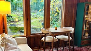 A cozy room with a wooden floor, a red patterned rug, a white sofa, a small table with chairs, and large windows overlooking a garden.