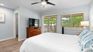 Ein Schlafzimmer mit Bett, Deckenventilator, Fernseher, Kommode und einem Fenster mit Blick auf Grünflächen.