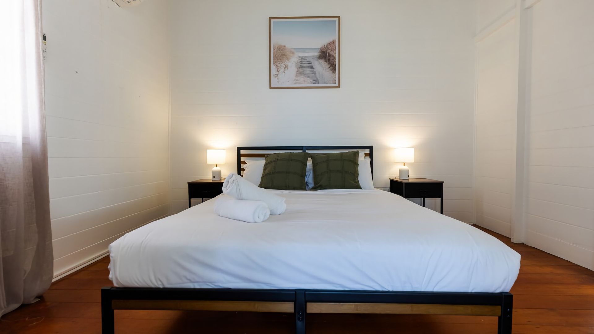 A neatly made bed with white linens and two pillows, a framed picture above the bed, and two bedside lamps on nightstands.