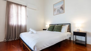 A neatly made bed with white linens and pillows, a bedside table with a lamp, and a painting on the wall. - Charming Cottage Stay (South Townsville)