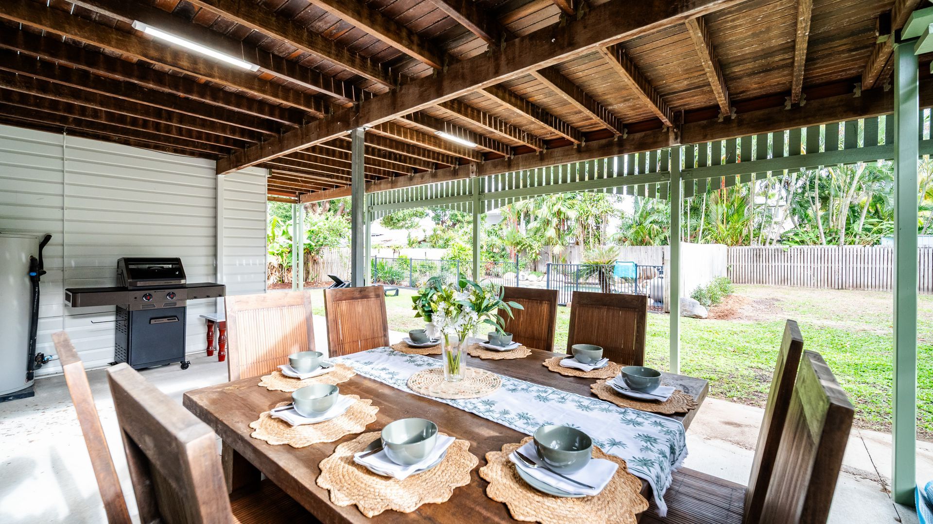 An outdoor covered patio with a wooden table set for dining, a grill, and a view of a garden.