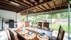 An outdoor covered patio with a wooden table set for dining, a grill, and a view of a garden. - Tropical getaway 10 min to city Vacation Home in Hermit Park (Hermit Park)