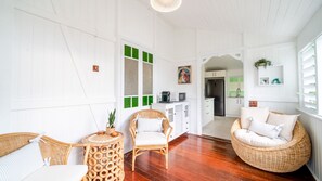 Quarto com piso de madeira, paredes brancas, uma cadeira de vime, uma pequena mesa redonda e uma área de cozinha ao fundo.