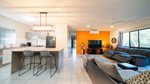 Een moderne keuken met een centraal eiland, een eethoek met barkrukken, een woonkamer met een bank en televisie, en een groot raam met uitzicht.