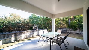 Een overdekt terras met een tafel en stoelen, omringd door een houten hek en bomen.