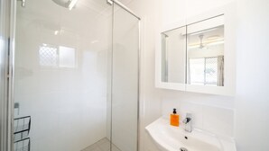 Een badkamer met een douchecabine van glas, een witte wastafel met een spiegel erboven en een zeepfles.