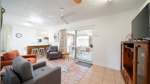 Una sala de estar con sofá, sillón y mueble para televisión. Se observa una zona de cocina con armarios blancos y una mesa de comedor.