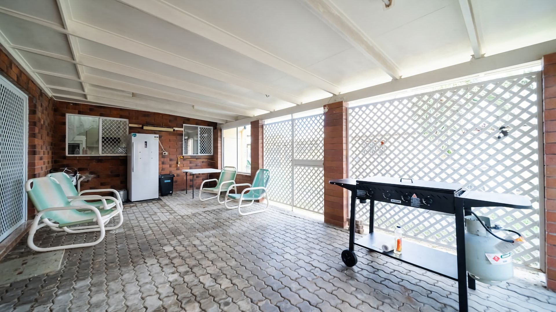A covered patio area with a brick wall, a refrigerator, two green chairs, a table, and a barbecue grill.