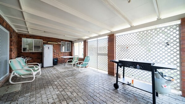 A covered patio area with a brick wall, a refrigerator, two green chairs, a table, and a barbecue grill.