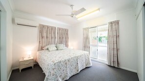 Un dormitorio con cama, mesita de noche, ventilador de techo y ventana con cortinas.
