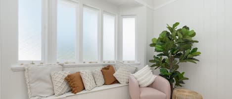 Ein Zimmer mit einer fest eingebauten Bank an einer weißen Wand, einem Blumentopf und einem Korbstuhl.