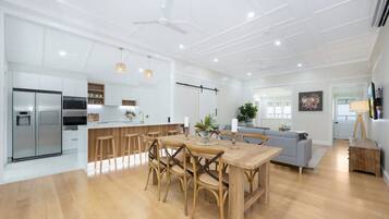 A modern kitchen with a built-in refrigerator, a dining area with a wooden table and chairs, and a grey sofa in a bright living space.