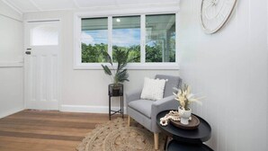 A room with a grey sofa, a small round table with a plant, a clock on the wall, and a window with a view of greenery. - Grand 4BR Queenslander on Harold – Sleeps 7 (West End)