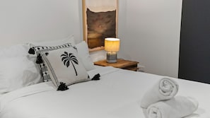 A neatly made bed with white bedding and decorative pillows, a bedside lamp, and a wall art piece. - Coastal Comfort Near The Strand (North Ward)
