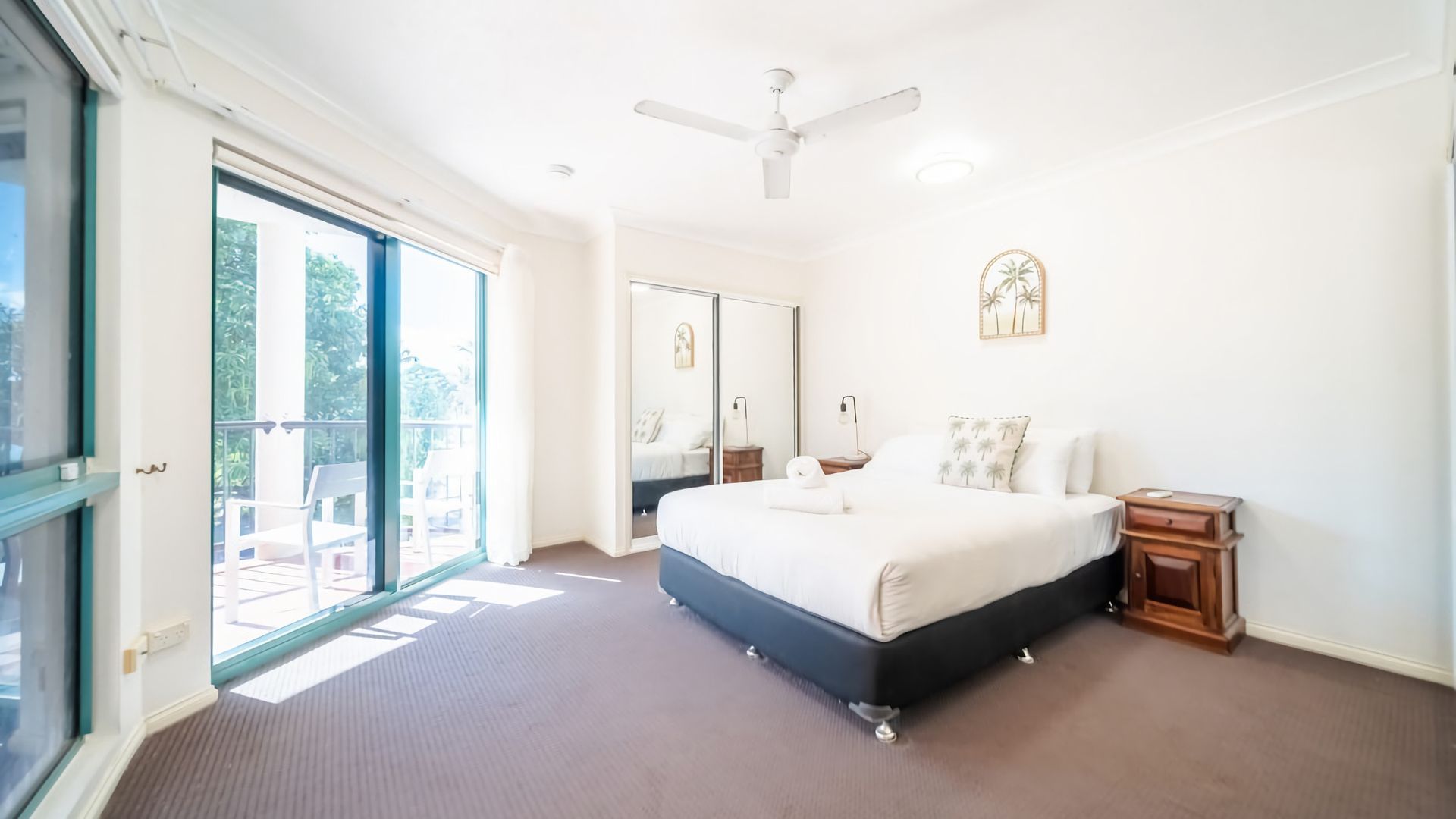 A bedroom with a large bed, a ceiling fan, a sliding glass door leading to a balcony, and a small bedside table.