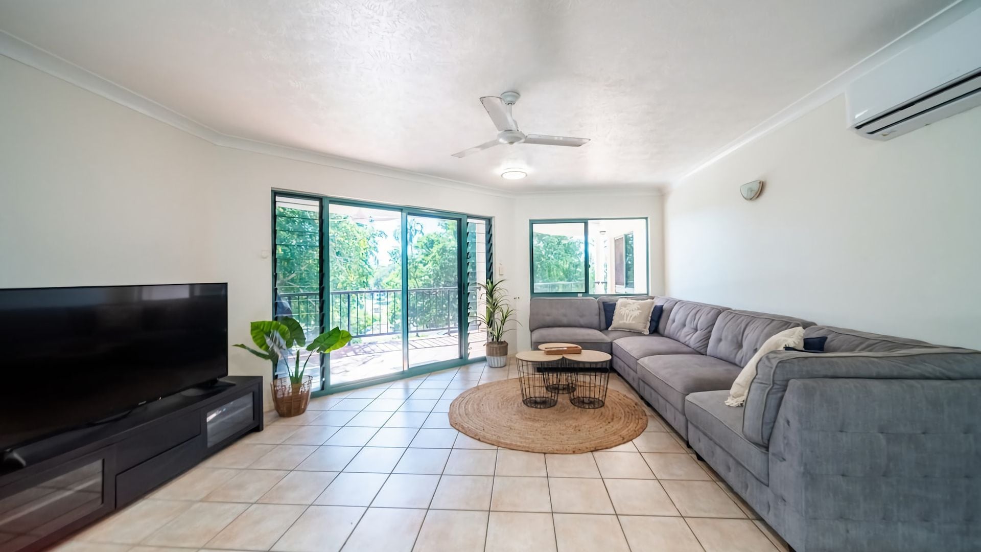 A living room with a flat-screen TV, a grey sofa, a coffee table, and a ceiling fan.