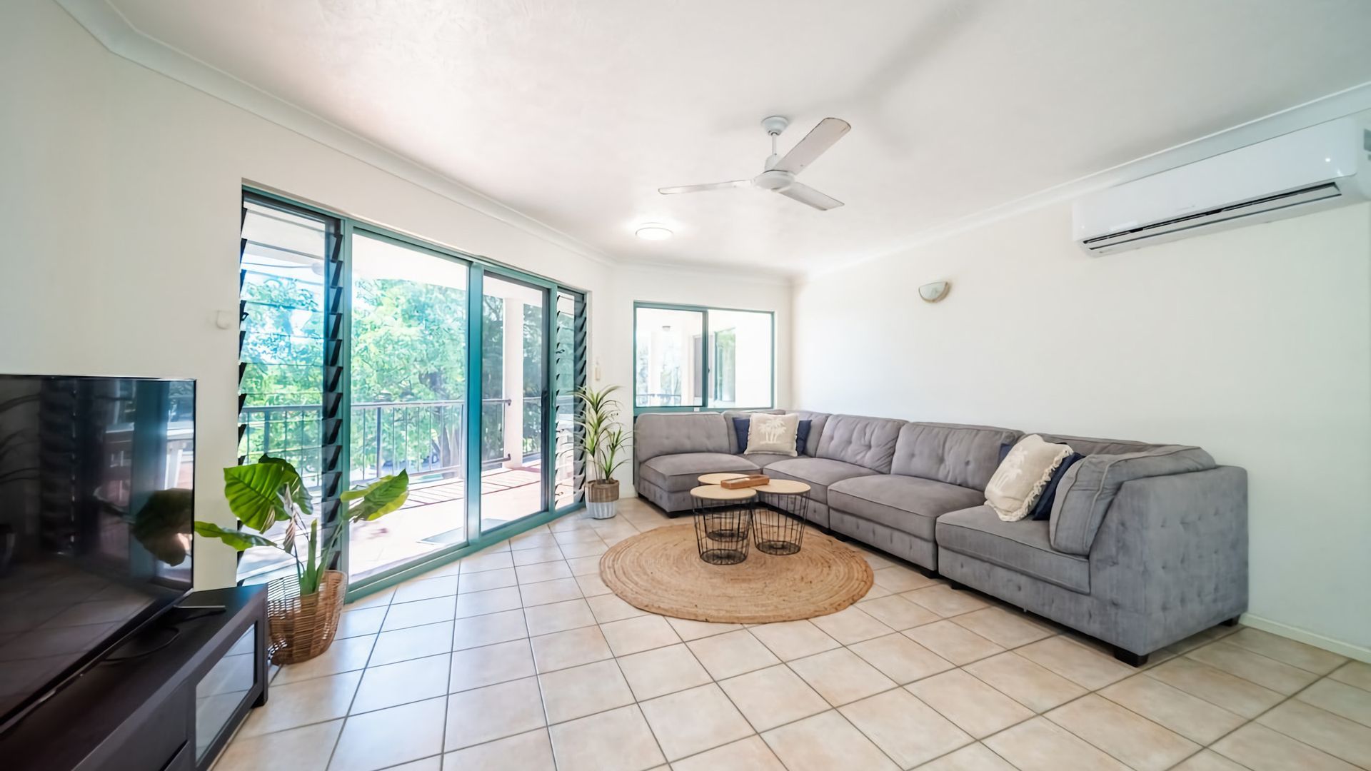 Ein geräumiges Wohnzimmer mit einem großen, grauen Ecksofa, einem Flachbildfernseher, einem Deckenventilator und einer großen Schiebeglastür, die auf einen Balkon im Freien führt.