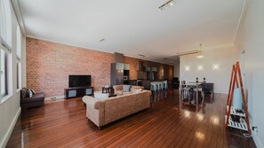 A spacious living area with a brick wall, wooden flooring, a sofa, a TV, and a kitchen area with bar stools. - Flinders Manhatten Residence (Townsville City)