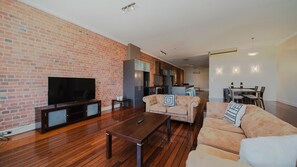 A modern living room with a brick wall, a flat-screen TV, a wooden coffee table, and a sofa with decorative pillows. - Flinders Manhatten Residence (Townsville City)