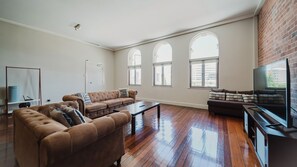 A spacious living room with a brown sectional sofa, a coffee table, and a flat-screen TV. There are large windows providing natural light and a view of the city. - Flinders Manhatten Residence (Townsville City)