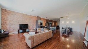 A modern living room with a brick wall, a flat-screen TV, a sofa, and a dining area with a kitchen in the background. - Flinders Manhatten Residence (Townsville City)