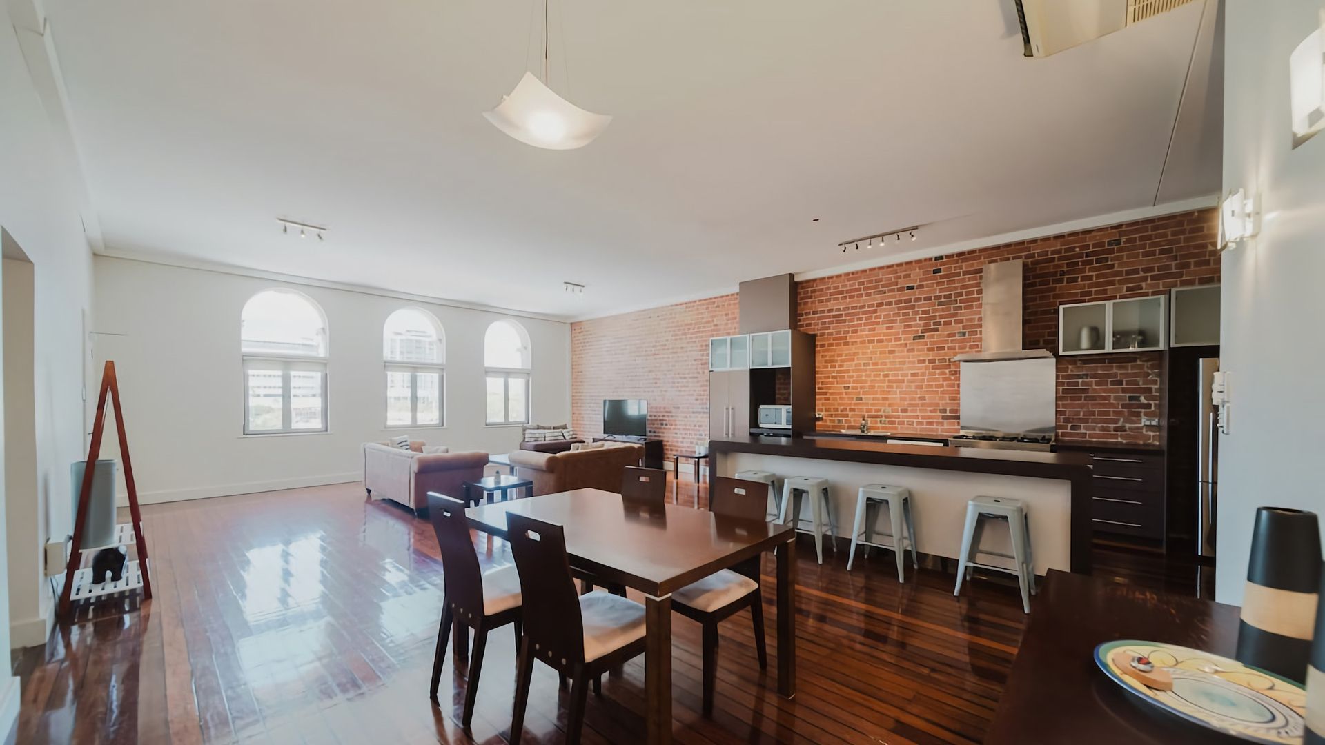 A spacious living area with a dining table, kitchen area with a brick wall, and large windows.