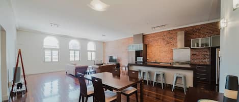 A spacious living area with a dining table, kitchen area with a brick wall, and large windows.