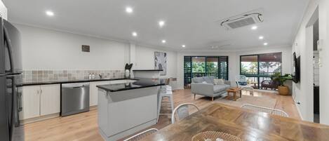 Une cuisine moderne avec un îlot central, un coin repas doté d’une table en bois, et un salon avec un canapé et un balcon offrant une vue dégagée.