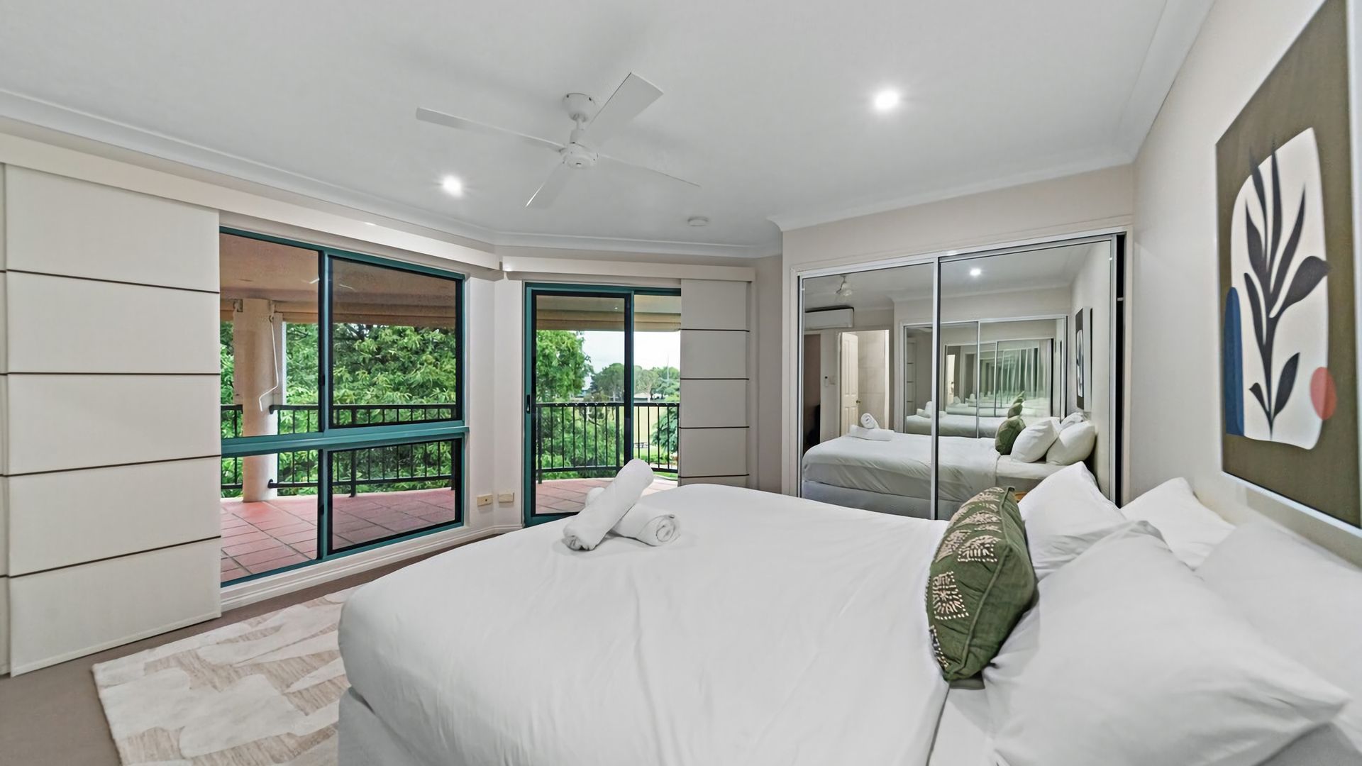 A bedroom with a large bed, a ceiling fan, a balcony with glass doors, and a view of greenery.