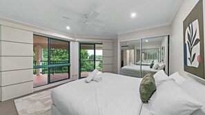 Een slaapkamer met een groot bed, een plafondventilator, een balkon met glazen deuren en uitzicht op groen.