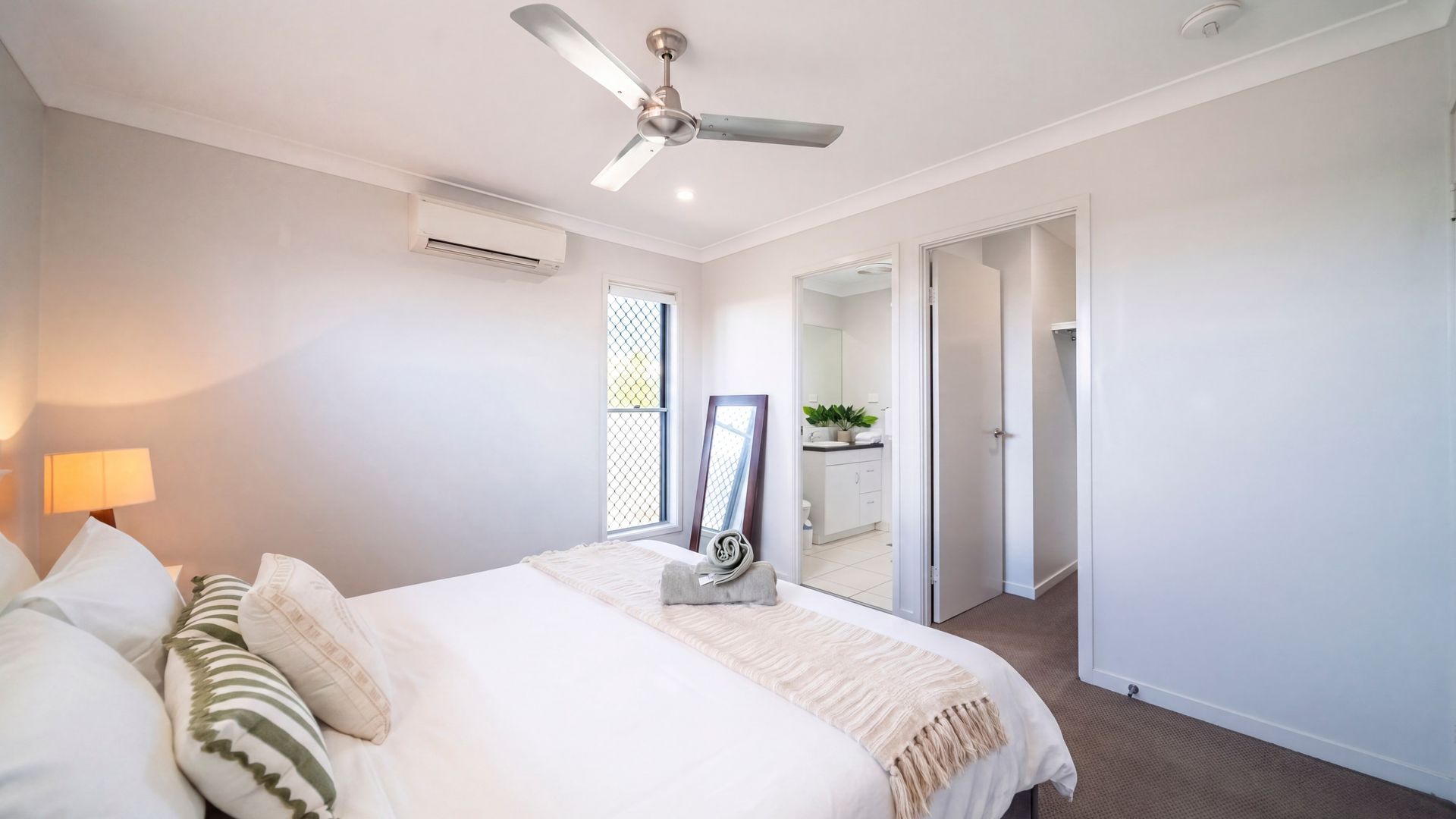 Een slaapkamer met een bed, een plafondventilator, een airconditioning en een raam met uitzicht, plus een badkamer die door een geopende deur zichtbaar is.