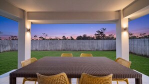 Eine überdachte Terrasse im Freien mit Tisch und Stühlen, ein Holzzaun im Hintergrund und ein klarer Himmel.