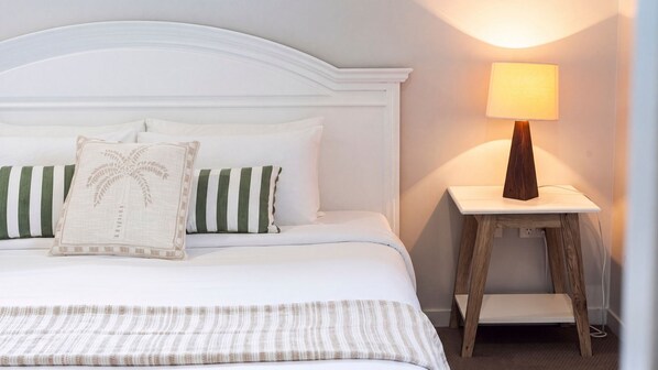 Een keurig opgemaakt bed met wit beddengoed en gestreepte kussens, een nachtkastje met een lamp en een houten nachtkastje.