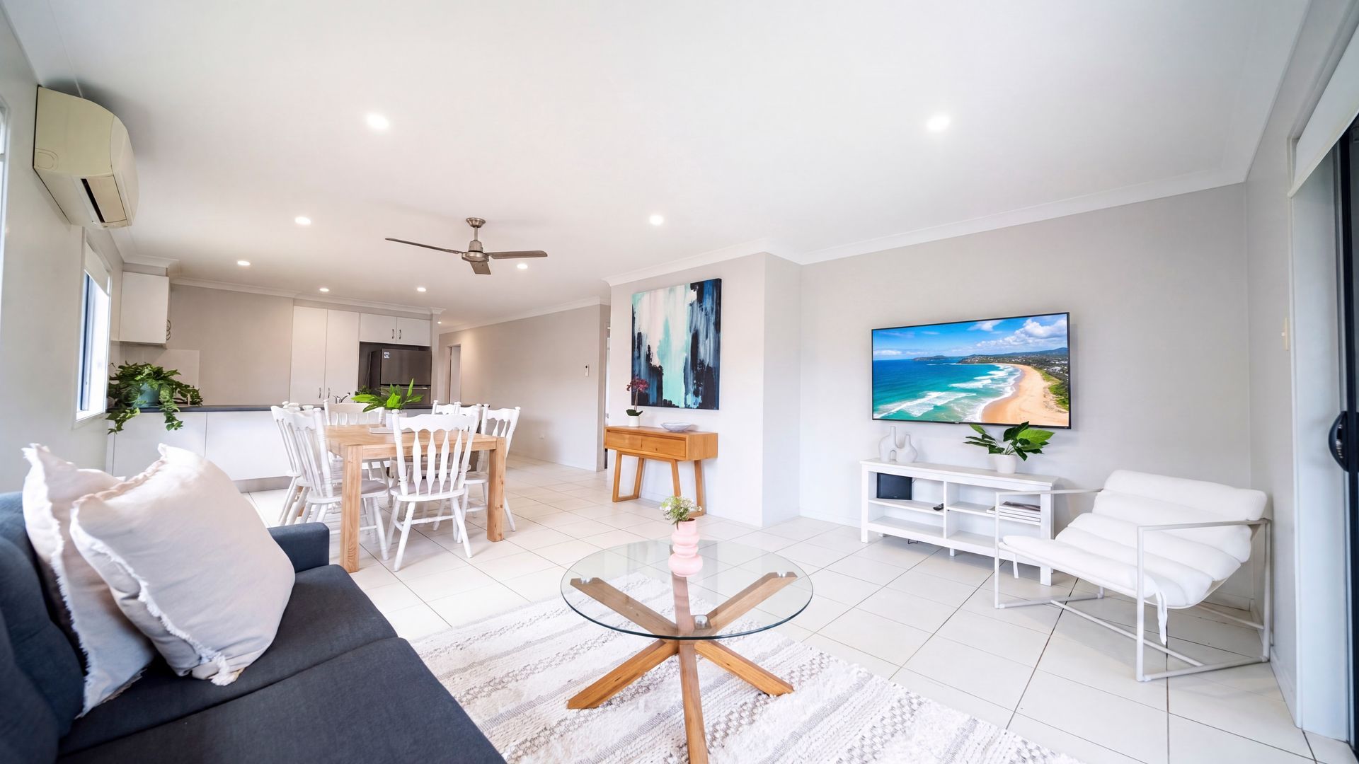 Ein modernes Wohnzimmer mit einer Couch, einem Glastisch, einem Essbereich mit Stühlen, einer Küche im Hintergrund und einem Fernseher, der eine Strandansicht zeigt.