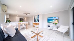 Een moderne woonkamer met een bank, een glazen salontafel, een eethoek met stoelen, een keuken op de achtergrond en een televisie waarop een zandstrand te zien is.