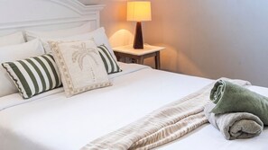 Een opgemaakt bed met kussens, een lamp en een opgerolde handdoek op een witte spreidek.