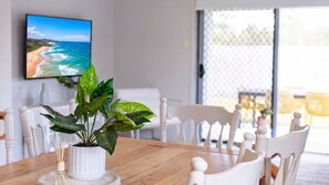 A dining area with a wooden table, white chairs, a potted plant, and a framed picture of a beach. - The Northside Haven (Burdell)