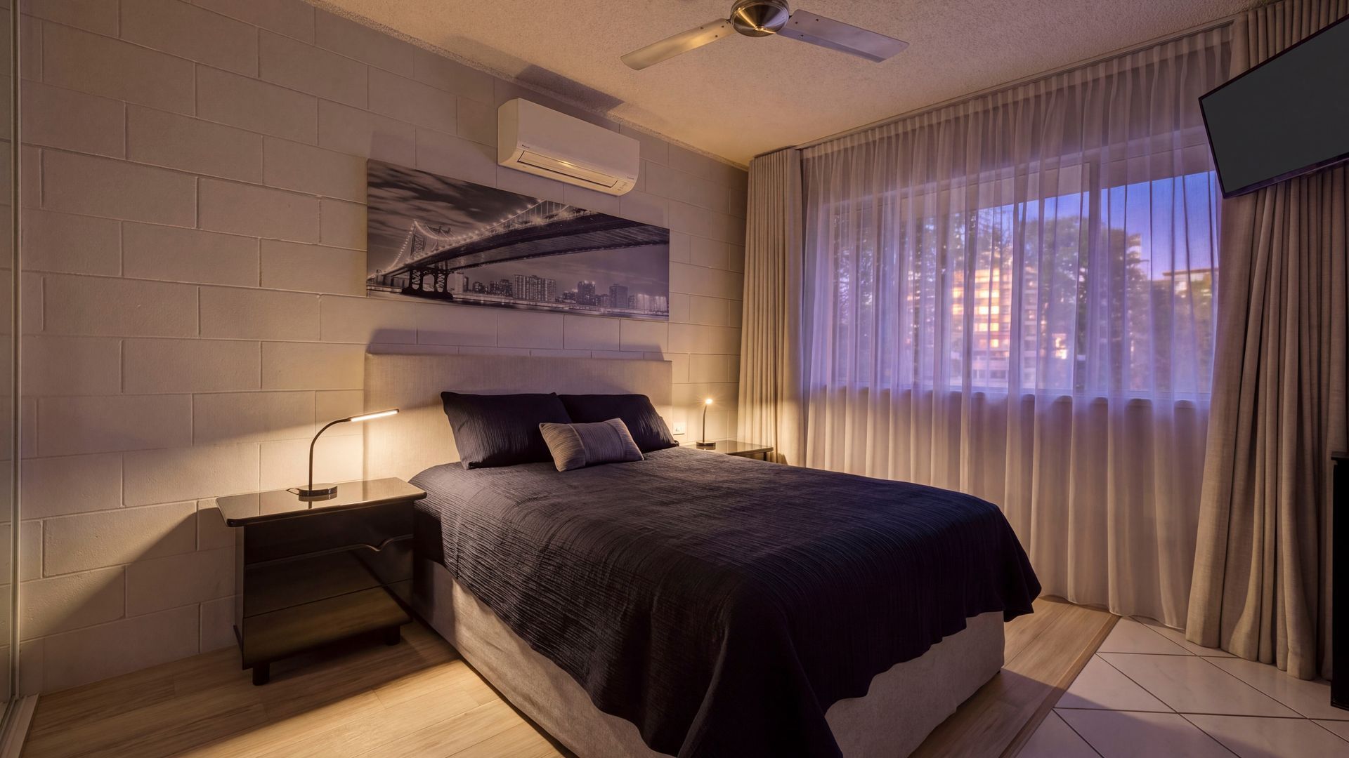 A modern bedroom with a bed, nightstands, a ceiling fan, and a cityscape artwork on the wall.