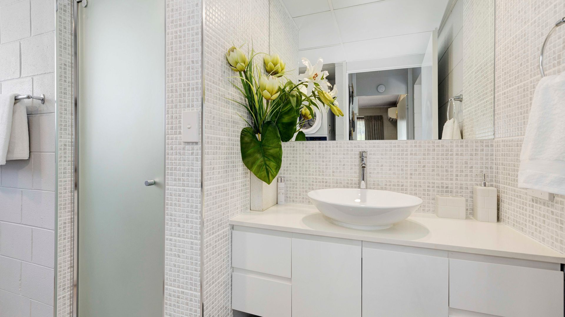A modern bathroom with a white sink, a large mirror, and a vase of flowers.