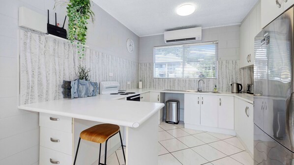 A modern kitchen with white cabinetry, a stainless steel refrigerator, a built-in oven, and a window with blinds. - Strand Oceanview Retreat (North Ward)