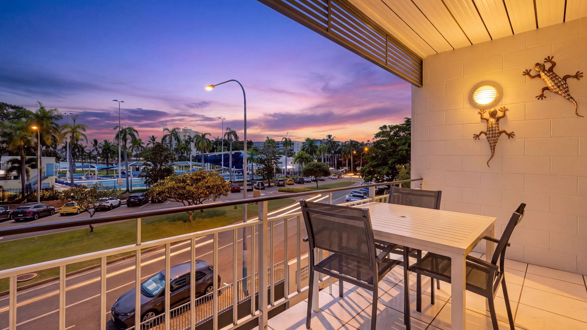 A balcony with a table and chairs, overlooking a street with cars and palm trees, and a sunset in the background.