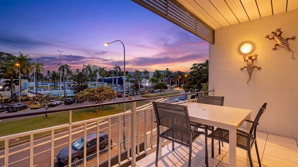 A balcony with a table and chairs, overlooking a street with cars and palm trees, and a sunset in the background.