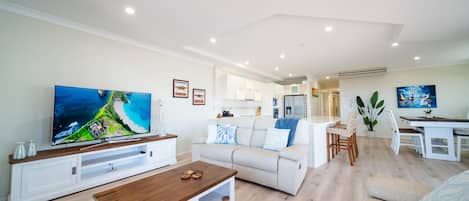 A modern living room with a flat-screen TV, a wooden coffee table, a white sofa, and a dining area with a white table and chairs.