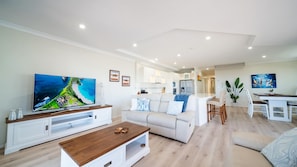 A modern living room with a flat-screen TV, a wooden coffee table, a white sofa, and a dining area with a white table and chairs.