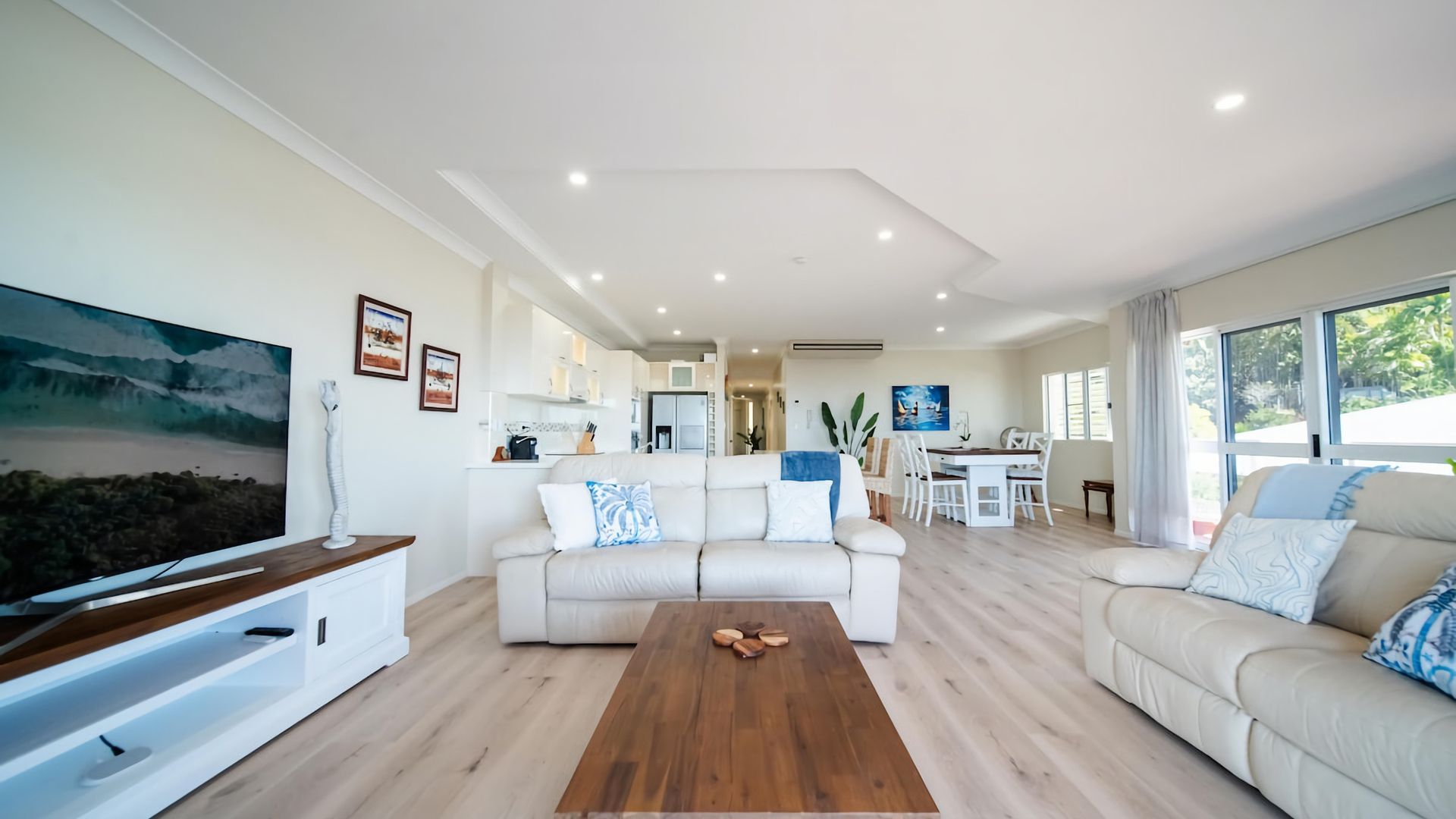 A modern living room with a sofa, coffee table, and a TV. There is a kitchen in the background with a dining area.