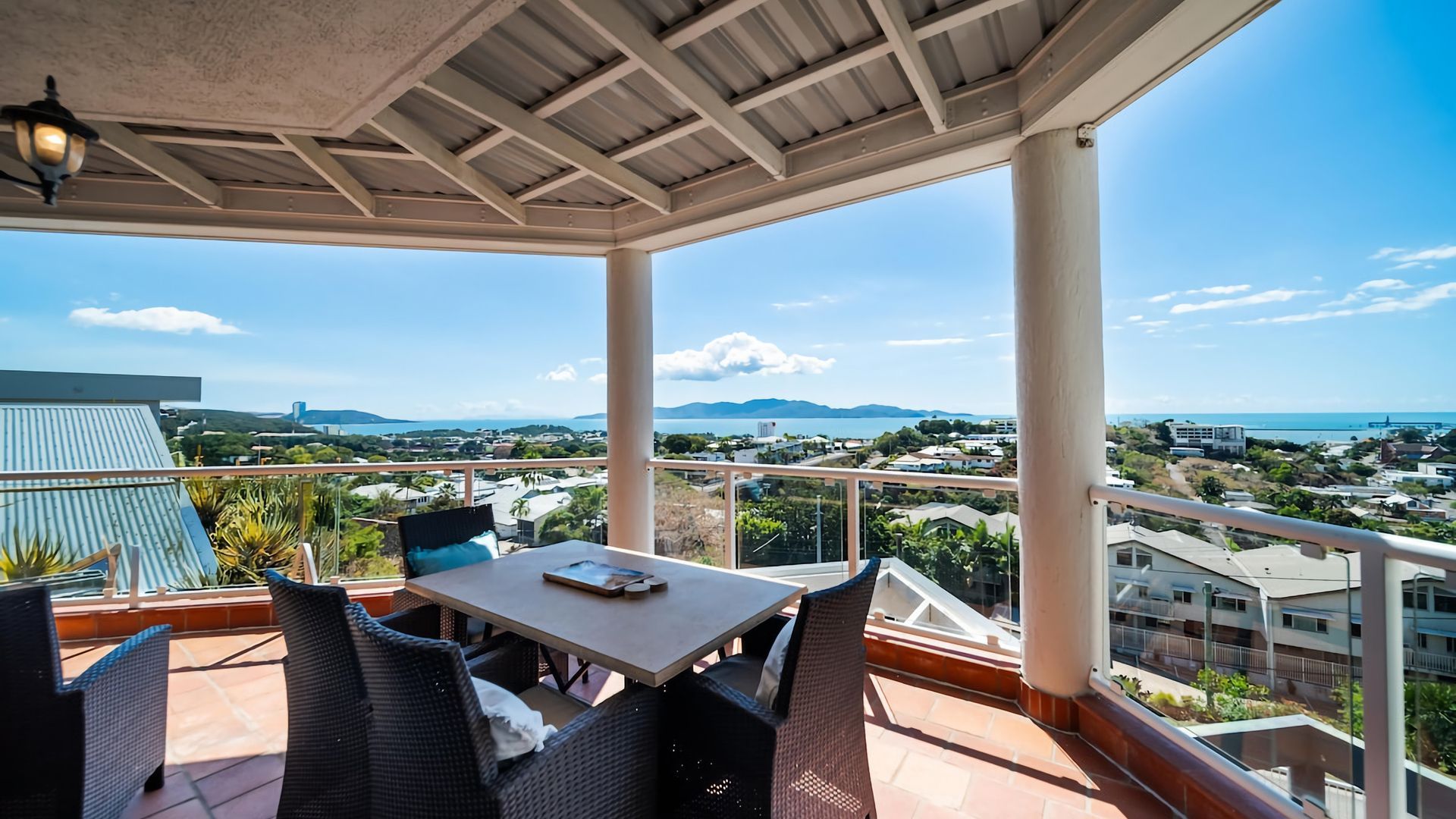 An outdoor patio with a table and chairs, overlooking a coastal landscape with buildings and mountains.