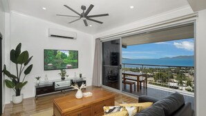 A modern living room with a large TV, a ceiling fan, and a view of the sea. - Searenity on Castle hill (Castle Hill)