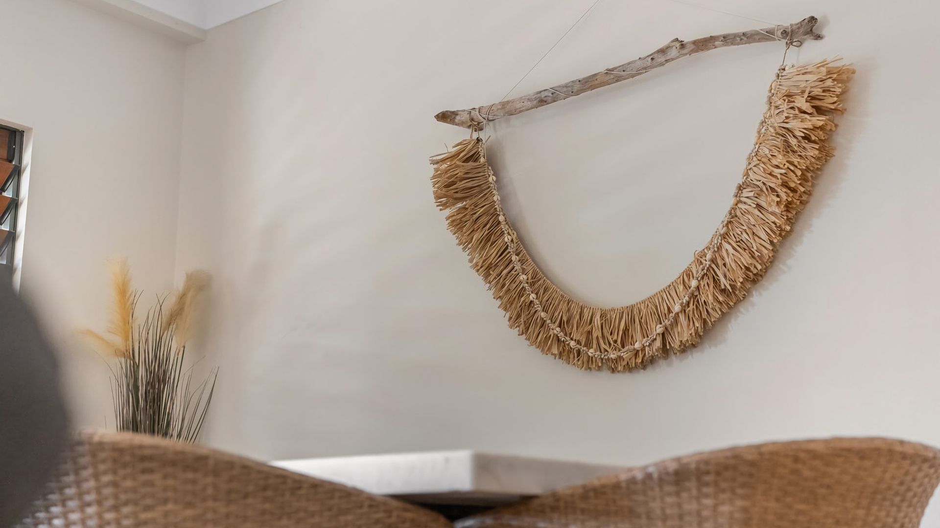 A room with a woven wall hanging, a wicker chair, and a vase with dried plants.