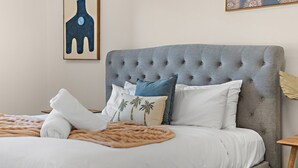Ein Bett mit einem gepolsterten Kopfteil, weißer Bettwäsche und dekorativen Kissen, darunter eines mit Palmenmotiven.